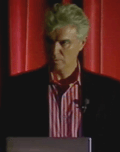Small picture of David Byrne behind a lectern, looking to the left.  Half his face is in shadow.