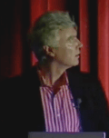 Small picture of David Byrne behind a lectern. He's looking up at the PowerPoint presentation behind him.
